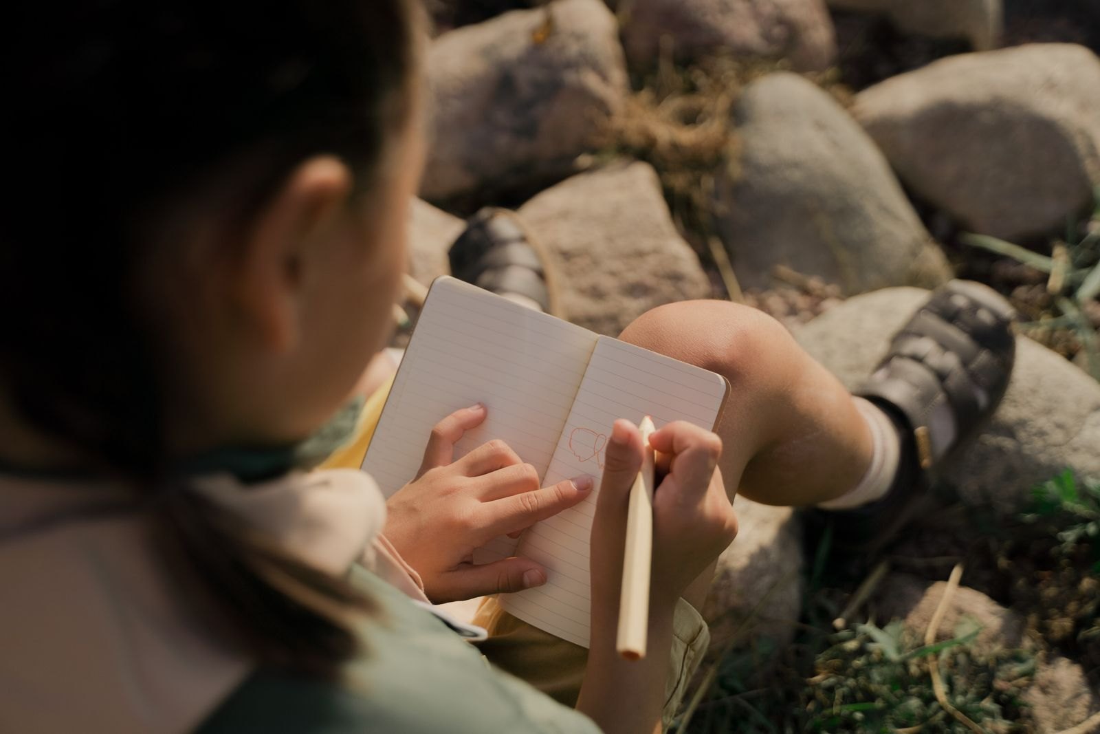 A kid sitting on rocks while writing on a notebook